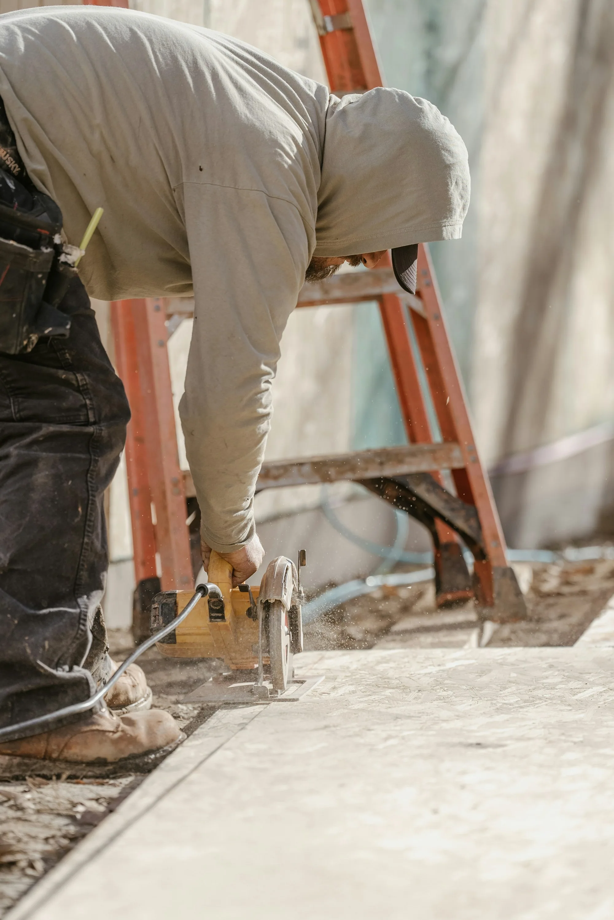 Carpenter cutting a board on site, with a worn tool belt and ladder nearby